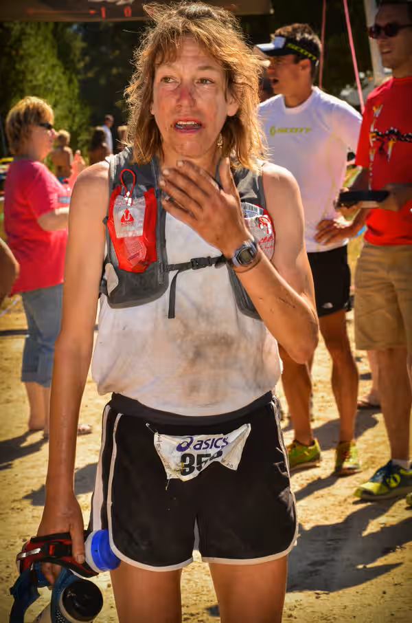 Ann Trason, exausta mas sorridente, cruza a linha de chegada de uma ultramaratona, com o rosto marcado pelo esforço e os braços erguidos em comemoração à conquista