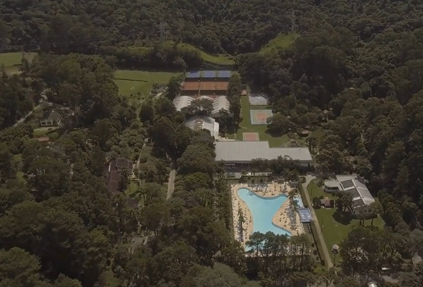 Vista aérea do Jaraguá Clube Campestre, com áreas de lazer, quadras de tênis e uma piscina em forma de lagoa, cercada por uma densa vegetação verde, criando um ambiente tranquilo e natural.