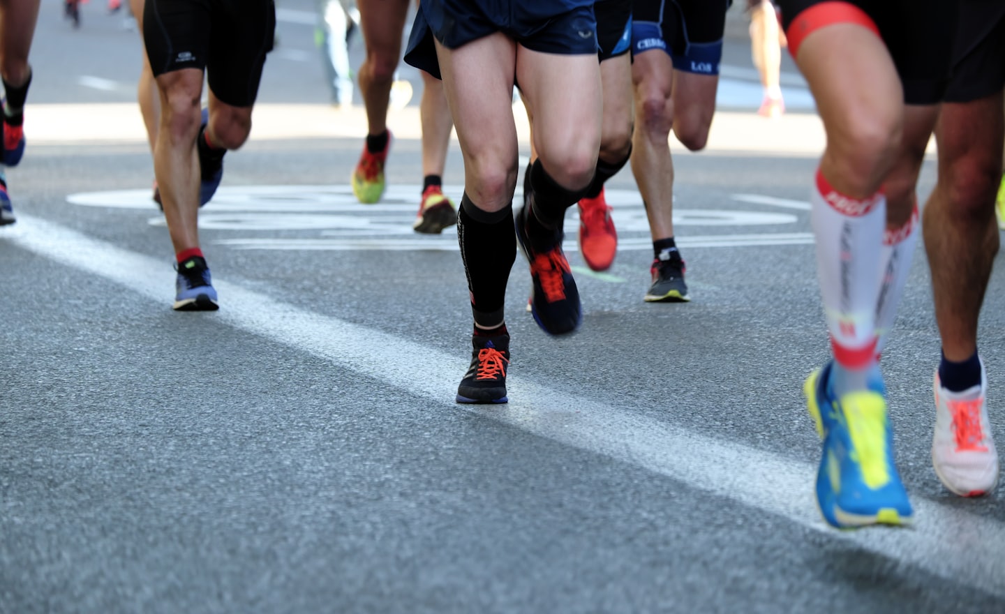 Pés de atletas calçados com tênis esportivos, em movimento rápido sobre o asfalto de uma rua durante uma corrida.